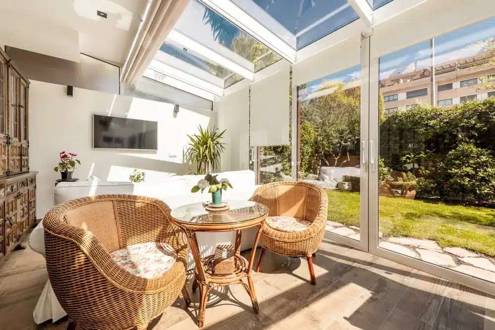 Intérieur d'une terrasse fermée en aluminium blanc et en verre avec de grandes portes coulissantes fermées donnant accès à un jardin avec pelouse et plantes ornementales, mobilier en bois et osier à l'intérieur, sol en grès.