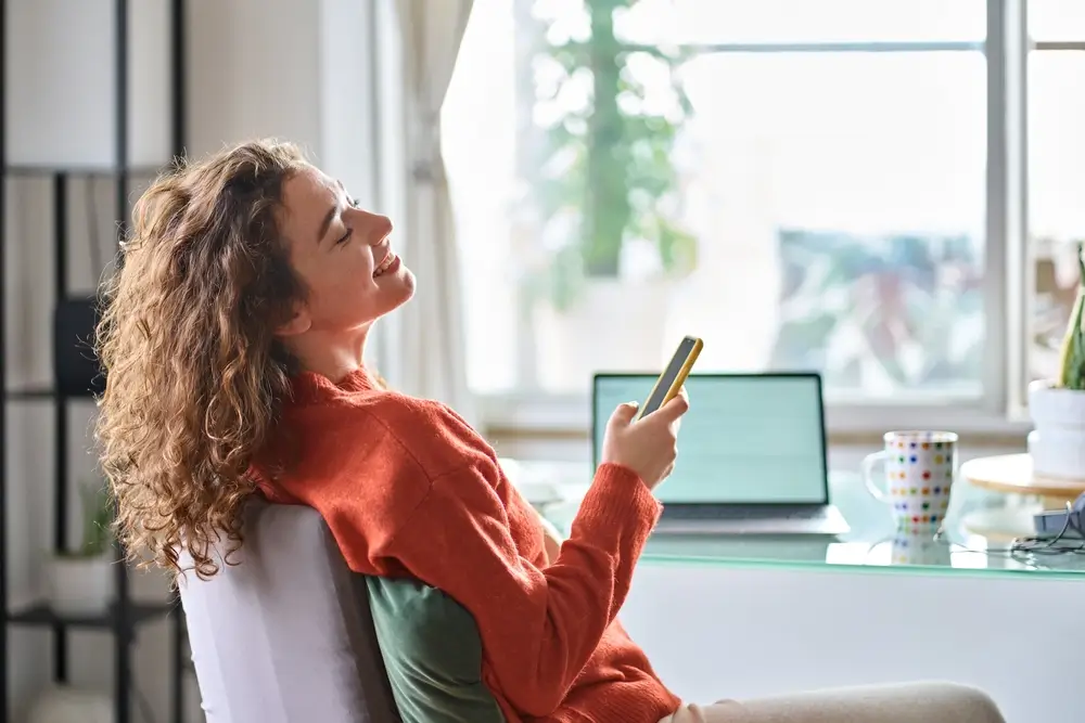 Jeune femme souriante et jolie tenant un smartphone utilisant un téléphone portable prenant une pause détente pendant qu'elle travaille à distance ou apprend de la maison assise sur une chaise à table avec un ordinateur portable.