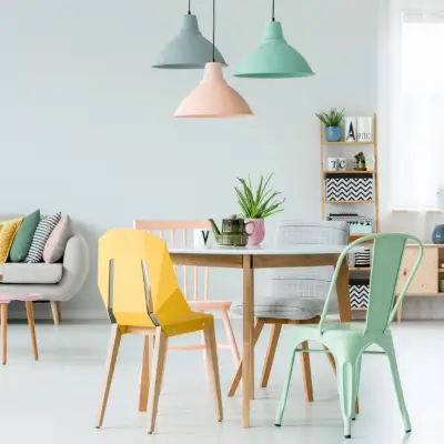Chaises modernes et colorées autour d'une table à manger sous des lampes pastel dans un salon avec des coussins sur le canapé adossé à un mur orné d'une affiche.