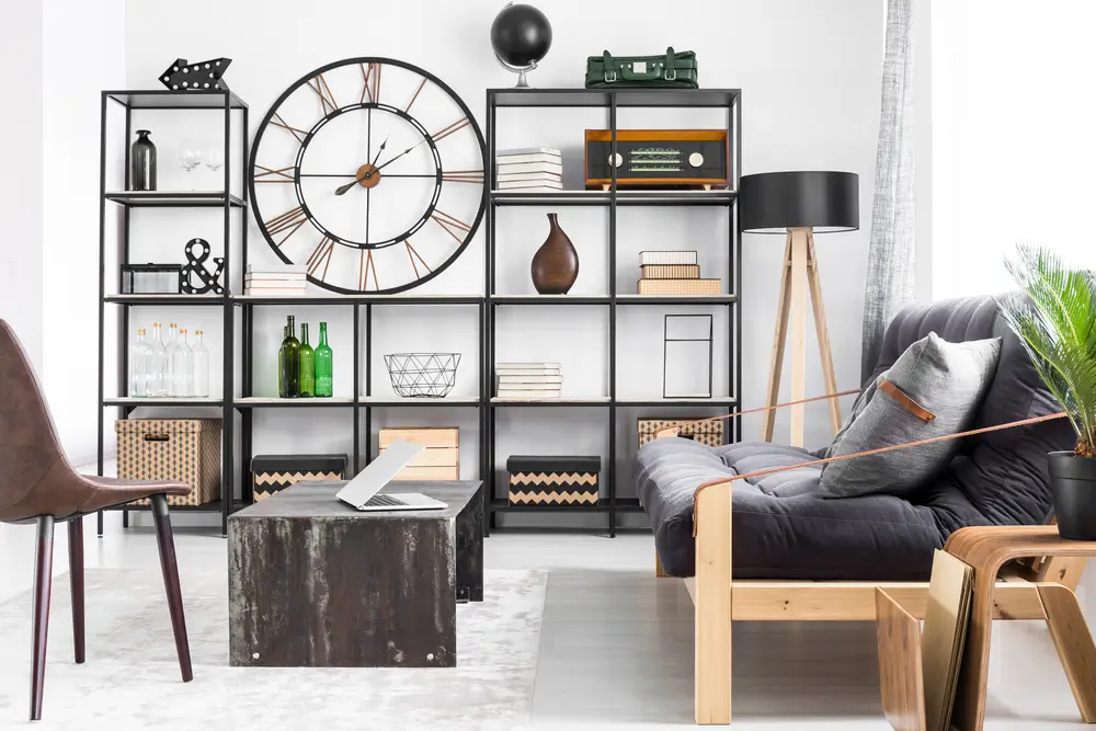 Laptop on table and grey pillow on couch in man's living room interior design with metal clock on the wall between shelves.